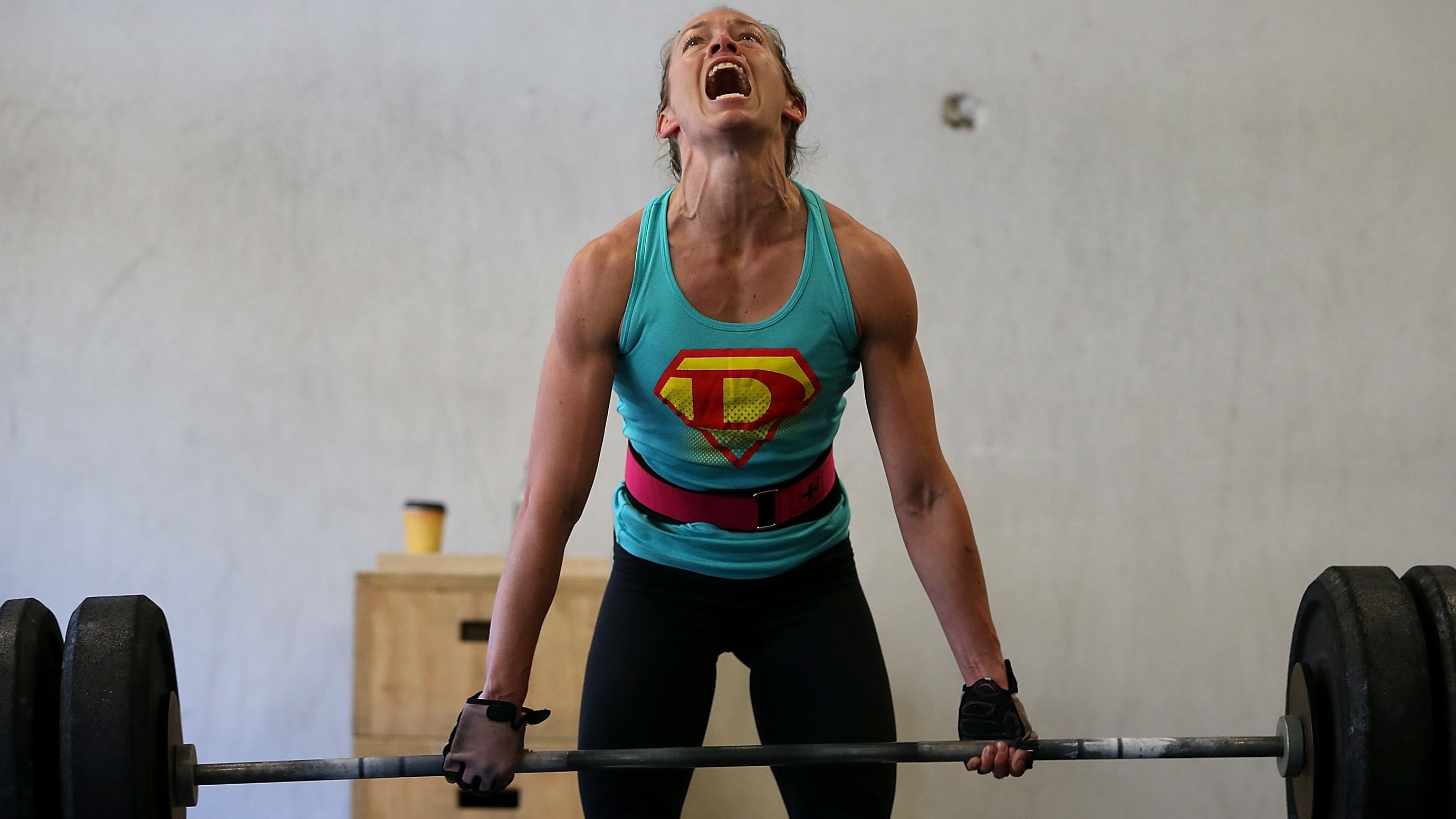 This One Exercise Can Slash Your Risk Of 8 Major Diseases Person in a turquoise tank top with a large “P” emblem and a pink lifting belt deadlifting a heavily loaded barbell in a gym, mouth open and head tilted back.