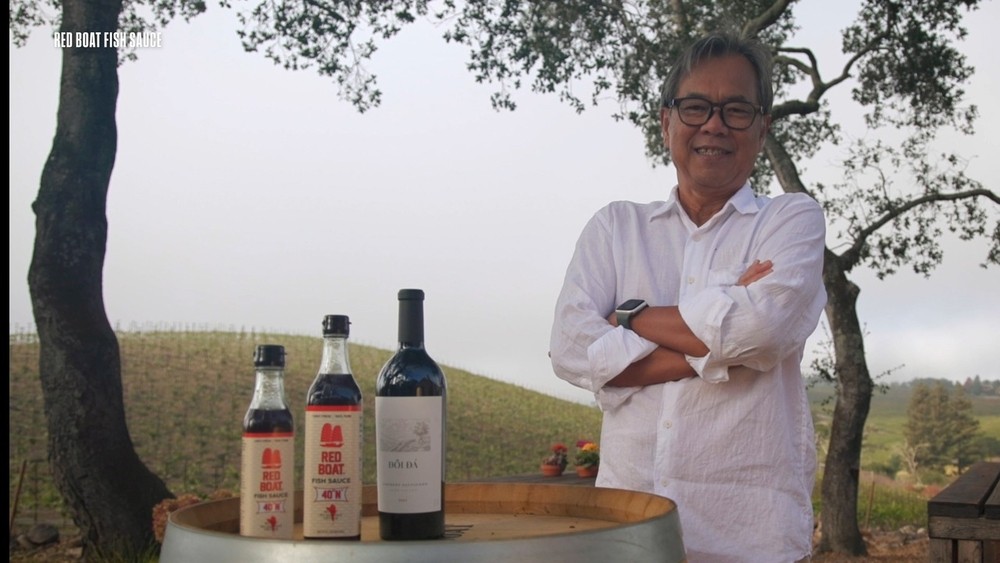Man in a white shirt standing with arms crossed beside two bottles of Red Boat Fish Sauce and a wine bottle on a barrel outdoors, with trees and hills in the background and the text “RED BOAT FISH SAUCE” in the top left.