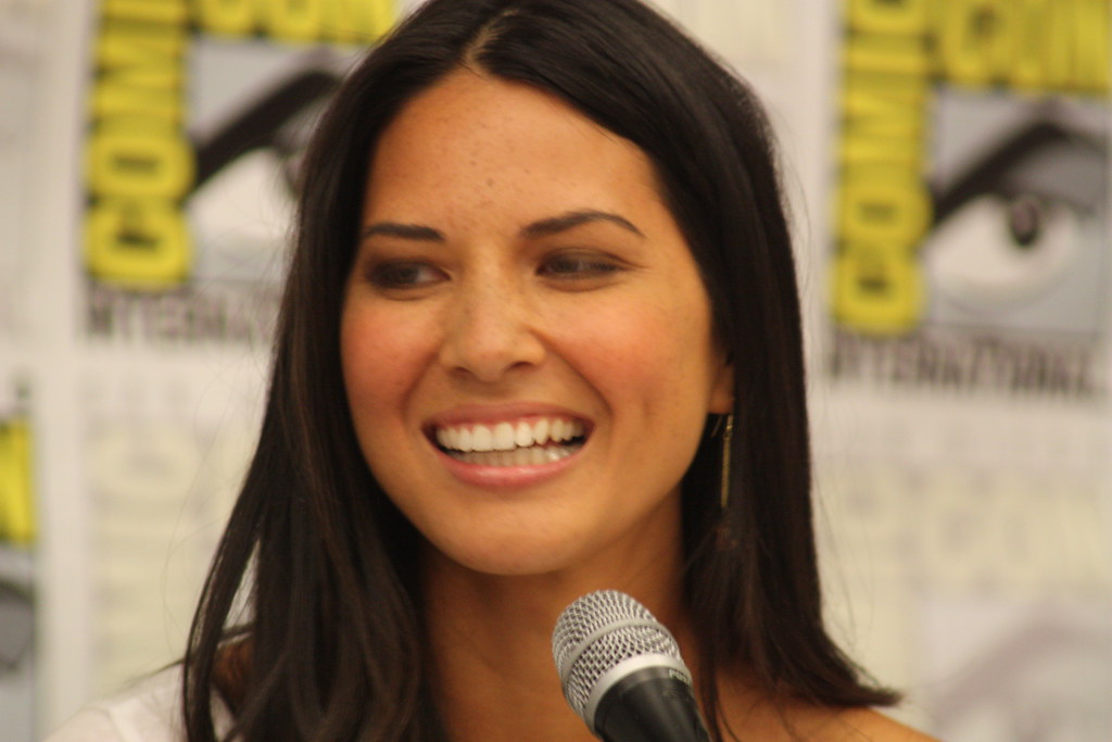 Olivia Munn Shares Powerful Moment She Finally Felt Beautiful After Cancer Battles Woman with long dark hair speaking into a microphone in front of Comic-Con International posters