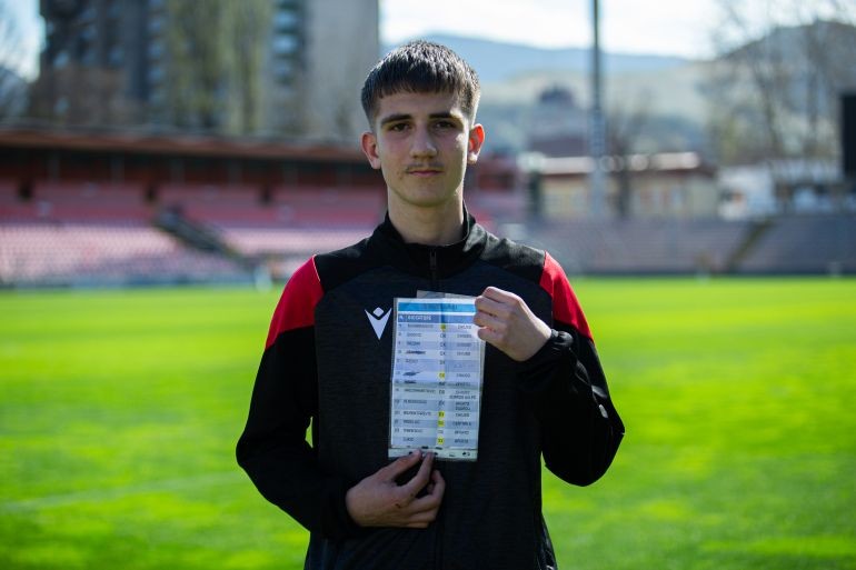 Bosnian Ball Boy Becomes Hero After Cleverly Foiling Italian Goalkeeper’s Plans Young man in a black and red tracksuit standing on a soccer field, holding up a card with rows of names and numbers; caption below reads “Bosnian Ball Boy Becomes Hero After Cleverly Foiling Italian Goalkeeper's Plans”