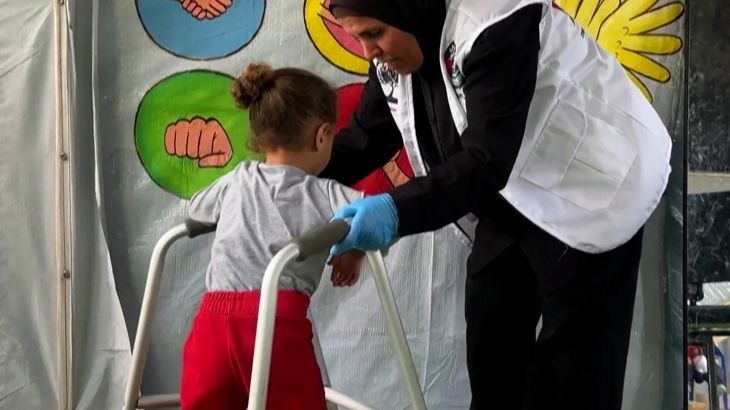 This Unique Gaza Camp Brings Joy and Safety to Children Woman in a white vest and blue gloves helping a child use a walker indoors, with a colorful wall behind them showing hand illustrations and the text, “This Unique Gaza Camp Brings Joy and Safety to Children”