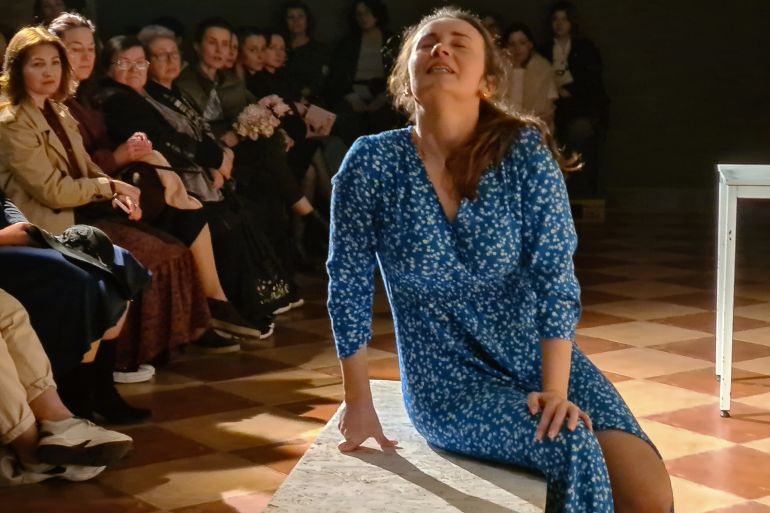 How Ukraine’s Veterans Are Transforming War Wounds Into Powerful Theatre Woman in a blue floral dress kneeling on a stage floor during a performance, with seated audience members in the background; text at the bottom reads, “How Ukraine's Veterans Are Transforming War Wounds Into Powerful Theatre”