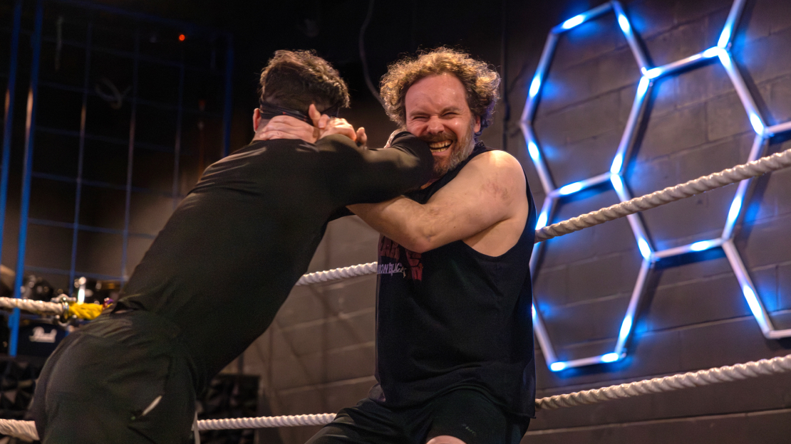 Meet The English Professor Teaching Storytelling Through Professional Wrestling Two men grappling in a wrestling ring, with one applying a hold near the ropes and blue hexagon lights on the wall behind them; text reads, "Meet The English Professor Teaching Storytelling Through Professional Wrestling"