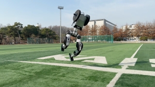 Meet The New Home Robot That Cooks, Cleans, And Organizes Your Life Effortlessly Humanoid robot running on a marked artificial turf field with buildings and trees in the background