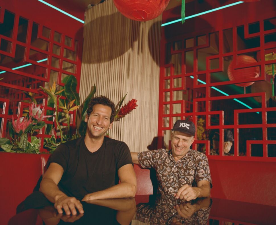 Genghis Cohen’s Beloved Chinese-American Flavors Make a Bold Move to Miami Two men sitting at a glossy table in a red restaurant interior with lattice screens, hanging lanterns, and tropical flowers; text reads, “Genghis Cohen's Beloved Chinese-American Flavors Make a Bold Move to Miami”