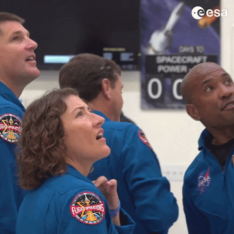 Artemis II Astronauts Revive Heartwarming Apollo Tradition in Space Four astronauts in blue flight suits look upward indoors; one sleeve patch reads “Flight Operations,” a NASA logo is visible, the ESA logo appears at top right, and text at the bottom reads “Artemis II Astronauts Revive Heartwarming Apollo Tradition in Space”