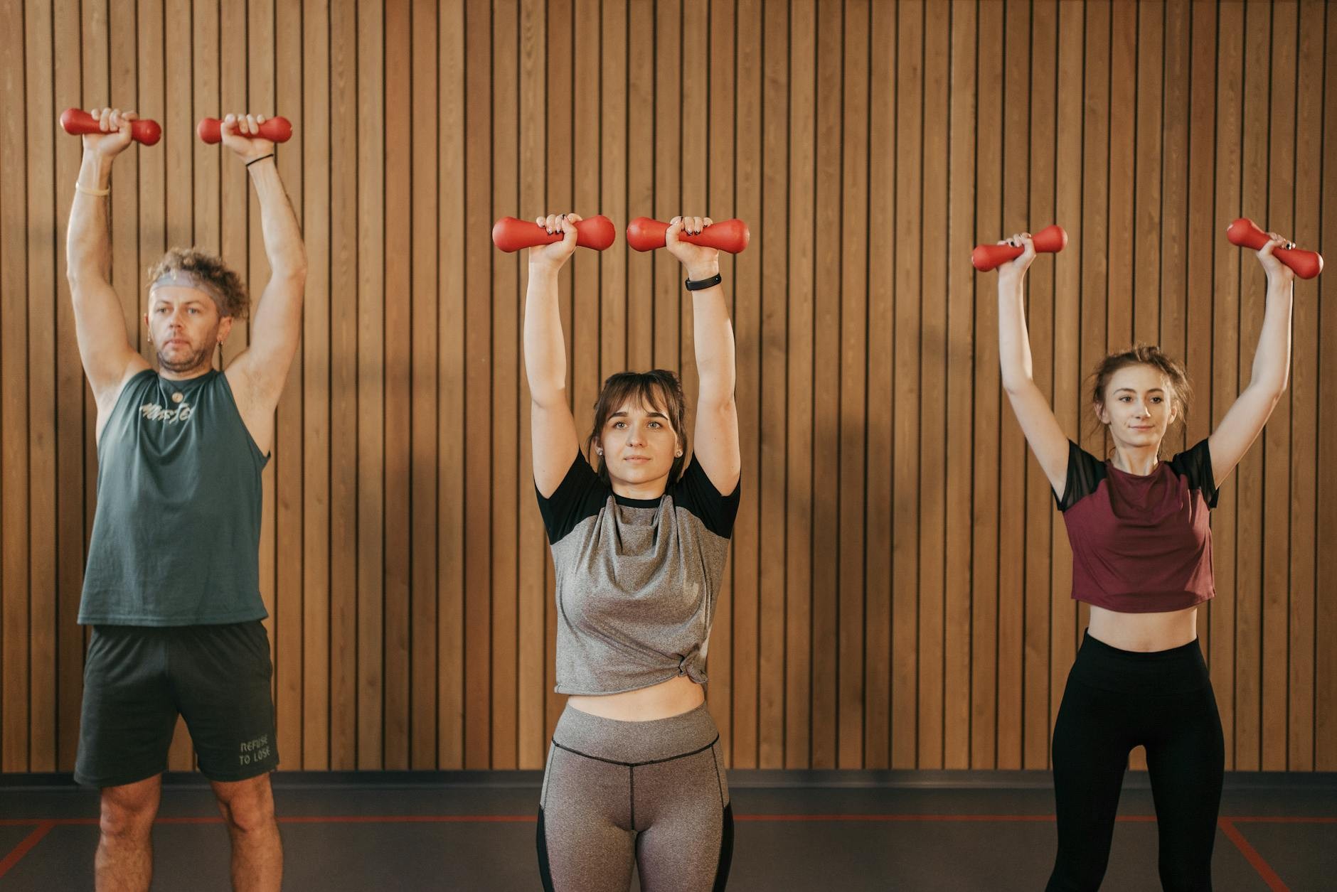These Simple Lifestyle Changes Could Slash Dementia Risk by 25% Three people in workout clothes raise red dumbbells overhead in front of a wood-paneled wall; text reads, “These Simple Lifestyle Changes Could Slash Dementia Risk by 25%”